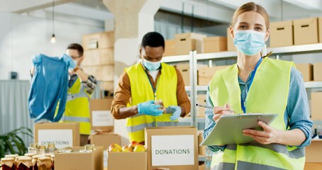 Wall Mural - Portrait of young Caucasian happy female volunteer in medical mask and uniform writing and smiling to camera. Multi-ethnic charity workers work in charitable stock organization preparing donations box