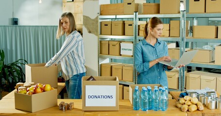 Wall Mural - Caucasian young beautiful woman charity center owner speaking with senior female worker in warehouse and typing on laptop. Woman preparing and packing donation box, social help, charity concept