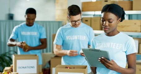 Wall Mural - Portrait of beautiful young concentrated African American woman volunteer standing in charity organization warehouse writing and checking donations. Male workers sorting donated stuff in boxes behind