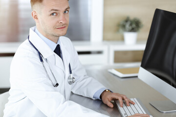 Doctor man sitting behind a computer at workplace in sunny clinic.