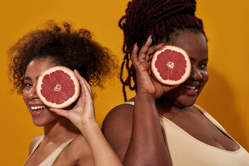 Wall Mural - Close up of two joyful african american women in underwear smiling at camera, covering one eye with a half of juicy grapefruit, posing isolated over orange background