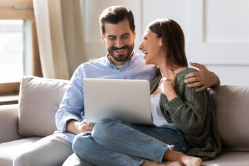 Wall Mural - Overjoyed millennial couple relax at home have fun laugh browsing internet on modern computer gadget. Smiling young Caucasian man and woman enjoy weekend use laptop together. Technology concept.