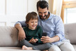 © fizkes - Smiling young Caucasian father and little son sit relax on couch using modern smartphone gadget play together. Happy dad and small boy child browse cellphone talk speak on webcam video call on device.
