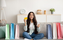 Positive Indian Woman With Credit Card And Smartphone Sitting On Sofa Surrounded By Shopping Bags At Home
