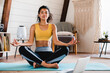 © InsideCreativeHouse - Beautiful young afro woman meditating on the floor at home. Home Training. Relaxed woman in practicing online sports, watching video tutorial on laptop computer, doing stretching yoga exercises.