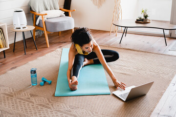 Young slim african girl stretching at home using laptop. Cheerful African woman stretching up In front of laptop, doing home workout, warming up for domestic fitness training, exercising 