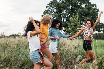 Wall Mural - Laughing woman runs in front of her friends with a raised hand. Group of four young females on summer vacation.