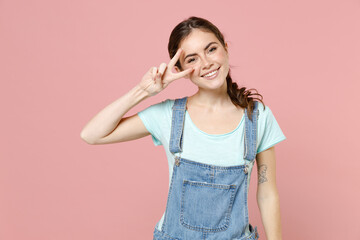 Wall Mural - Young friendly positive smiling caucasian woman in trendy denim clothes blue t-shirt cover eye with victory v-sign gesture isolated on pastel pink background studio portrait. People lifestyle concept