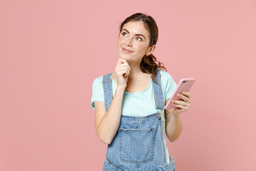 Wall Mural - Young wistful dreamful thoughtful caucasian woman 20s wear denim clothes blue t-shirt hold mobile cell phone look aside isolated on pastel pink background studio portrait. People lifestyle concept