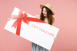 Young satisfied happy fun woman in summer clothes striped dress straw hat hold in hand large huge gift voucher flyer mock up looking camera isolated on pastel pink color background studio portrait