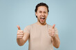 © ViDi Studio - Young smiling happy friendly cheerful caucasian attractive man 20s wear casual basic beige t-shirt show thumb up gesture isolated on pastel blue background studio portrait. People lifestyle concept