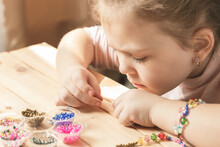 A Little Girl Is Engaged In Needlework, Making Jewelry With Her Own Hands, Stringing Multi-colored Beads On A Thread
