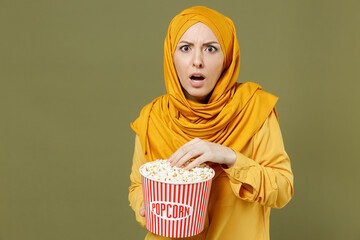 Poster - Young shocked confused arabian asian muslim woman 20s in abaya hijab yellow clothes takeaway pop corn bucket watching film movie isolated on olive green background People uae islam religious concept.