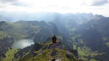 A Young Man Is Standing By Himself Alone On Top Of The Mountain At The Edge, Looking Down Over The Beautiful And Magical Landscape With A Small, Pretty Lake And Sunbeams Hitting The Lush Greenery.