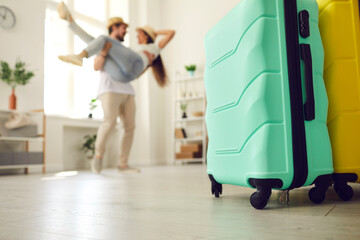 Ground level shot two suitcases of mint and yellow colors standing on floor in living-room with happy couple in love having fun in blurred background. Romantic travel, dream vacation, perfect holiday