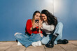 © Mikel Allica - Couple of friends with long hair, one blonde and one brunette, looking at their cell phones sitting on the floor in front of a blue wall.