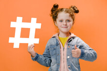 Happy Attractive Smiling Caucasian Little Girl Child Holding Big White Hashtag Symbol Showing Thumbs Up Gesture, Posing Isolated On Pastel Orange Studio Background. Like To Popular Blog And Trends