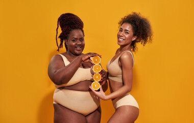 Wall Mural - Studio shot of cheerful african american women in underwear smiling while holding ripe juicy orange halves, posing isolated over orange background