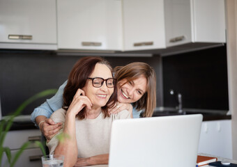 adult daughter and her mature mother looking at the laptop screen and laughing while making online s