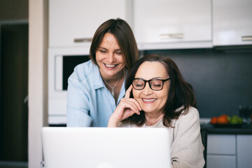 Adult daughter and her mature mother looking at the laptop screen and laughing while making online shopping. Smiling young woman teaching her parent how to use a computer.