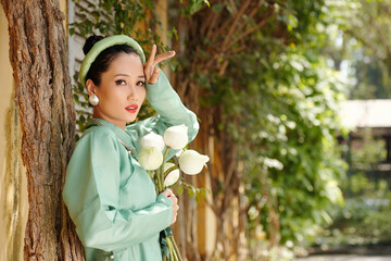 Poster - Portrait of serious young Vietnamese woman standing at tree with bouquet of lotus flowers in hands and looking at camera