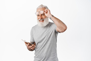 Happy old man with tattoos, take off glasses and using digital tablet, smiling at camera, standing in grey t-shirt against white background