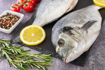 Fresh fish dorado on black slate cutting board and grey concrete background with ingredients for cooking