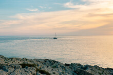 Sailboat Is Sailing Far Out In The Sea Against The Backdrop Of A Sunset. View From The Rocky Shore