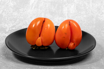 Wall Mural - Two ugly ripe orange persimmons on black plate on light grey textured backdrop close up.