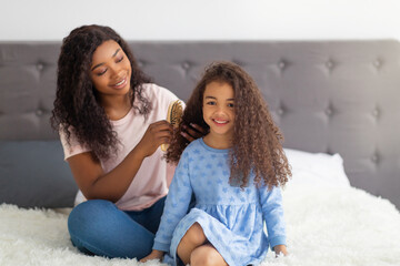 Canvas Print - Loving black mom brushing her pretty daughter's hair on bed at home