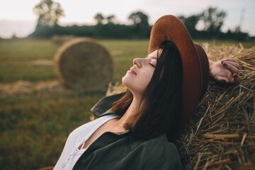 Beautiful stylish woman with herb in mouth relaxing on haystack in summer evening field. Tranquility