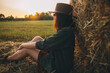Beautiful stylish woman in hat sitting at haystacks in evening sunset in summer field. Atmospheric