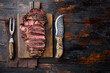 Grilled or fried and sliced marbled meat steak rib eye, on wooden serving board, with meat knife and fork, on old dark  wooden table background, top view flat lay, with copy space for text