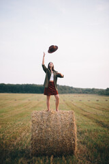 Wall Mural - Beautiful carefree woman throw hat up in the sky standing on haystack in summer field. Happiness