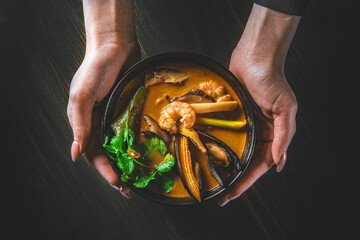 Poster - pan asian food. woman hands holding Tom Yam soup in black bowl on wooden table background