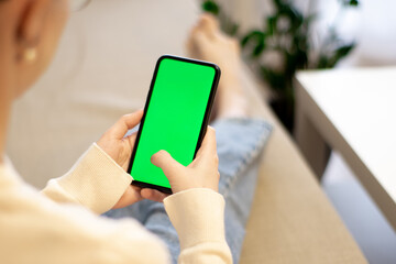 Woman at home, on the couch, using a smartphone with a green mockup screen. Girl using a mobile phone on the Internet. Point of view shot