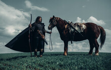 Woman In Image Of Medieval Warrior Stands Near Horse In Field.