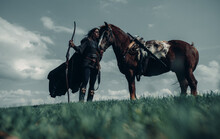 Woman In Image Of Medieval Warrior Stands Near Horse In Field.