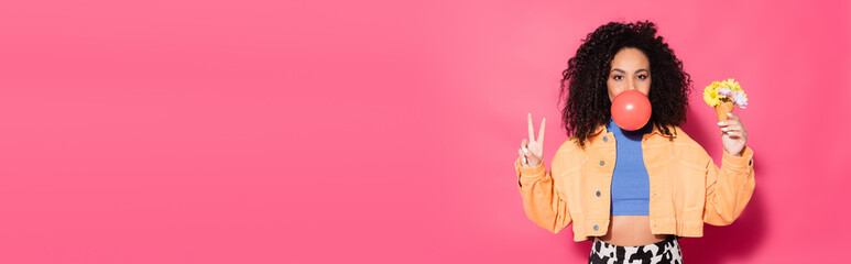 curly african american woman blowing bubble gum, holding waffle cone with flowers and showing peace sign on pink, banner