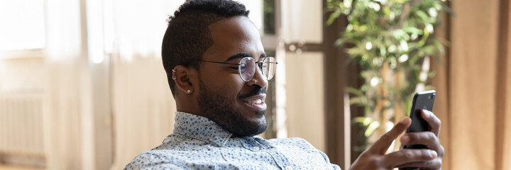 Happy millennial guy using online bank app on smartphone for payment. African man reading article on internet on cell phone, ordering on store website, making video call from home. Panorama photo