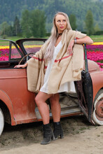 Young Pretty Blond Woman In White Summer Dress And Warm Scarf In Front Of Old Rustic Car On Colorful Tulip Fields. Chilliwack Tulip Festival. British Columbia. Canada 
