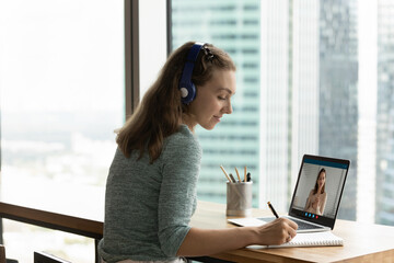 Young Caucasian woman in headphones talk speak on video zoom call with business partner make notes. Female colleagues have webcam digital online conference meeting on laptop. Virtual event concept.