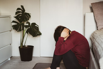 Depressed and sad woman covering her head with hands and sitting on the floor in the bedroom. Troubled with domestic violence, unwilling pregnancy, abortion, trolling. Mental crisis, depression