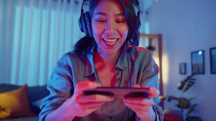 Wall Mural - Happy asia girl gamer wear headphone competition play video game online with smartphone colorful neon lights in living room at home. Esport streaming game online, Home quarantine activity concept.