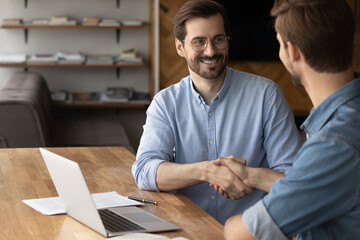 Happy young Caucasian businessman shake hand close deal or make agreement with client or colleague in office. Smiling male business partners handshake get acquainted greeting. Collaboration concept.