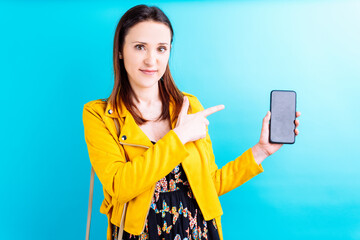 beautiful young woman in yellow jacket on blue background pointing and showing a smartphone