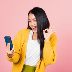 Poster - Happy Asian portrait beautiful cute young woman excited holding mobile phone and gesturing ok sign, studio shot isolated on pink background, Thai female making finger symbol on smartphone