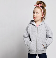 Portrait of naughty frolic kid girl in gray hoodie with zipper standing with hands in pockets, looking aside and sticking out her tongue over light background with copy space