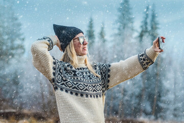 Outdoor close up portrait of young beautiful woman with long hair wearing hat, sweater posing with her phone in nature view. Christmas, winter holidays concept. Snowfall. Snow trees on background.
