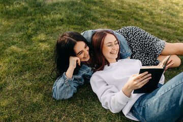 Two best friends spending time outdoors, lying on green grass in a park and reading a book, laughing.